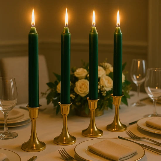 Four tall dark green altar candles in gold candlesticks, lit and arranged on a dining table with plates, wine glasses, and a floral centerpiece in the background.
