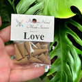 Crystal Fairy Patch Love premium hand-made cone incense in a clear plastic bag held against a green monstera leaf background