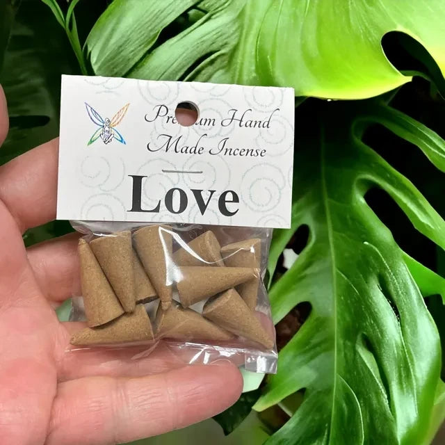 Crystal Fairy Patch Love premium hand-made cone incense in a clear plastic bag held against a green monstera leaf background