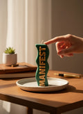 Crystal Fairy Patch green and beige wavy sculptural candle with raised MANIFEST lettering on a ceramic plate on a wooden desk.