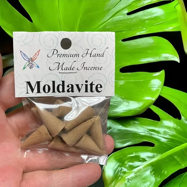 Crystal Fairy Patch Moldavite premium hand-made cone incense in a clear plastic bag held against a green monstera leaf background.