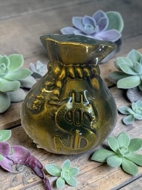 Crystal Fairy Patch handcrafted money bag candle with gold dusting and dollar sign emblem, surrounded by decorative succulents.